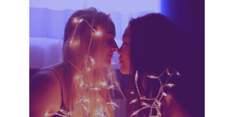 Image description: Two women facing each other. Their eyes are closed and their noses are touching. The woman on the left has long blonde hair and the woman on the right has long black hair. They are draped in fairy lights. (Image from Unsplash)