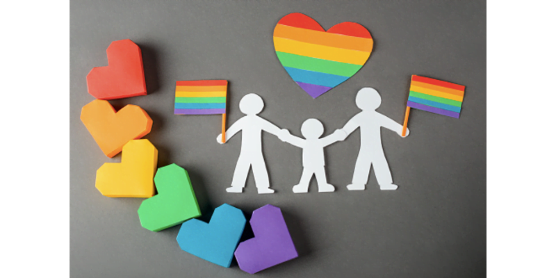 Image description: On a grey background, there are 3D paper hearts in rainbow colours on the left and two larger paper dolls holding hands with a smaller paper doll in the middle. The paper dolls are holding LGBTQ+ flags in their free hands and there is a rainbow striped heart above them.