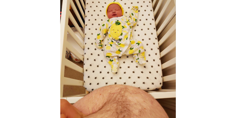Image Description: The photo is taken from a white birthing person's perspective, looking down past his now empty tummy to his baby lying in the cot.
His hairy tummy, which only a few hours before would have eclipsed this view, is now empty and he is pressing it in with his left hand to show the absence of both baby and organs.